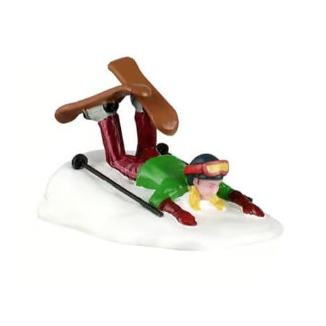 This image shows one of the figurines. It was designed to look like a girl who fell while skiing. She's laying face down in the snow with her skis in the air and the ski poles are in the snow. She has blonde hair and is wearing a blue hat with red goggles, a green jacket, red gloves and red pants. The skis are brown and the ski poles are black.