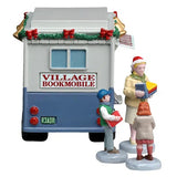 This image shows what this item looks like from the back. It reads "Village Bookmobile" and has garland decorated with bells and red bows. The license plate reads "R3ADR." The little boy is wearing a blue hat, a green and white jacket and jeans. The librarian is wearing a Santa hat, a yellow jacket, a yellow skirt and is holding a bunch of books. The little girl is wearing a light brown jacket and a pink dress.