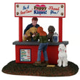 This image shows what this Lemax table accent looks like on a white background. It was designed to look like a booth set up in the grass. The sign on the booth reads "For a great cause! Puppy Kisses! Thank you!" There's a person behind the booth counter lifting dogs up for a woman to see. There's a sign on the booth with suggested donations: Wet Kiss $3. Sloppy Kiss $4. Dainty Kiss Free. 3 Dog Puppy Kiss $10. Biscuit $1. There's a white dog waiting patiently at the front of the booth.