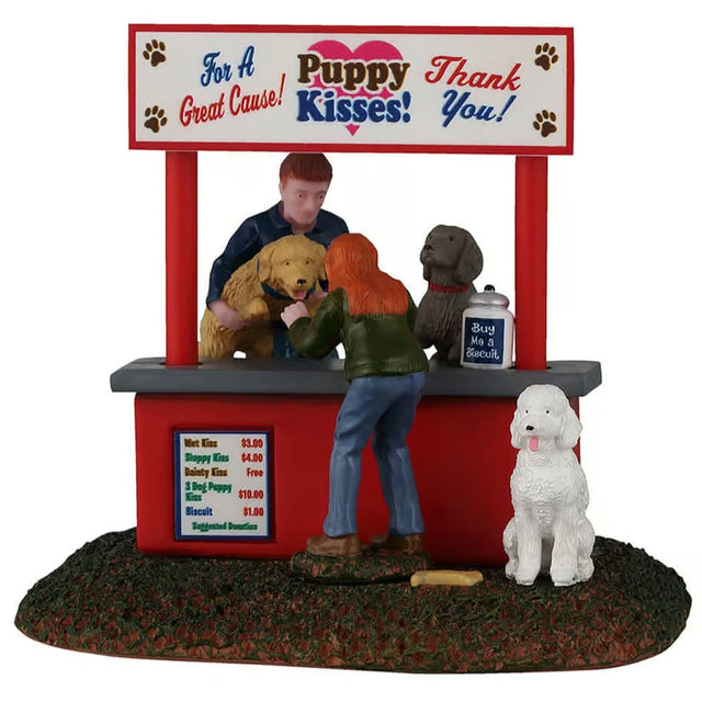 This image shows what this Lemax table accent looks like on a white background. It was designed to look like a booth set up in the grass. The sign on the booth reads "For a great cause! Puppy Kisses! Thank you!" There's a person behind the booth counter lifting dogs up for a woman to see. There's a sign on the booth with suggested donations: Wet Kiss $3. Sloppy Kiss $4. Dainty Kiss Free. 3 Dog Puppy Kiss $10. Biscuit $1. There's a white dog waiting patiently at the front of the booth.