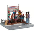 This image shows what this table piece from Lemax looks like on a white background. It was designed to look like a a group of people singing songs on a snow-covered stone sidewalk. There's a sign behind them that reads "Pine Valley Middle School Christmas Concert. December 24, 1-3pm. Everyone welcome!"