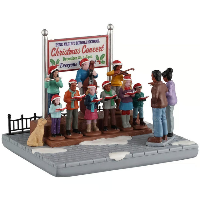 This image shows what this table piece from Lemax looks like on a white background. It was designed to look like a a group of people singing songs on a snow-covered stone sidewalk. There's a sign behind them that reads "Pine Valley Middle School Christmas Concert. December 24, 1-3pm. Everyone welcome!"
