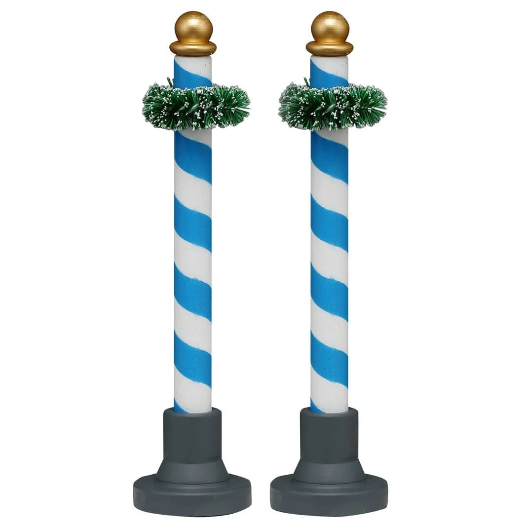 This image shows what these two Lemax Village accessories look like on a white background. These were designed to look like decorative poles. They're blue and white striped with a gold bauble on top. Near the top of the pole, there's a green garland wrapped around it.