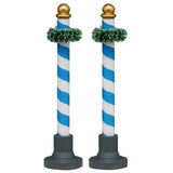 This image shows what these two Lemax Village accessories look like on a white background. These were designed to look like decorative poles. They're blue and white striped with a gold bauble on top. Near the top of the pole, there's a green garland wrapped around it.