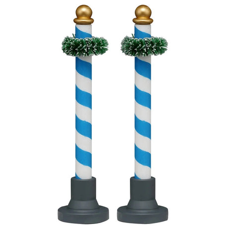 This image shows what these two Lemax Village accessories look like on a white background. These were designed to look like decorative poles. They're blue and white striped with a gold bauble on top. Near the top of the pole, there's a green garland wrapped around it.