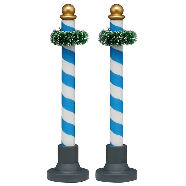 This image shows what these two Lemax Village accessories look like on a white background. These were designed to look like decorative poles. They're blue and white striped with a gold bauble on top. Near the top of the pole, there's a green garland wrapped around it.