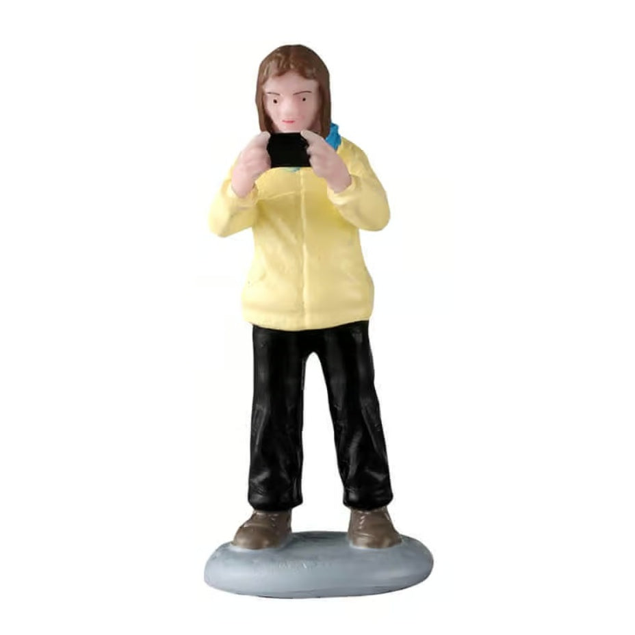 This figurine was designed to look like a woman taking a picture on her phone. She has brown hair and is wearing a yellow jacket, black pants and brown shoes.