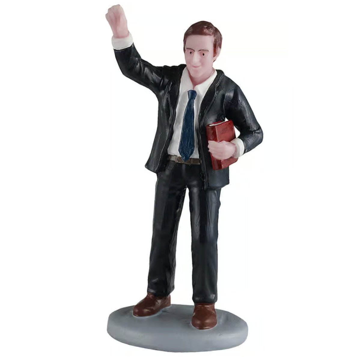 This image shows what this Lemax General Collectibles figurine looks like on a white background. It was designed to look like a young man with brown hair, wearing a black suit with a dark blue tie. He's waving with one hand and holding a Bible with the other.