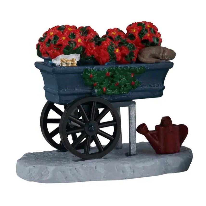 This figurine was designed to look like a cart with two wheels that has a bunch of holly flowers inside it. There's a brown watering jug on the ground underneath it.