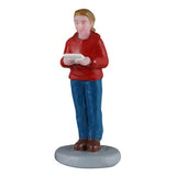 This image shows a young girl figurine holding a sheet of music and singing. She has light brown hair and is wearing a red hoodie, blue jeans and brown shoes.
