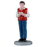 This image shows a man figurine holding a sheet of music and singing. He has brown hair and is wearing a white long sleeve shirt with a red vest, blue jeans and black shoes.