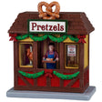 This image shows what this Lemax table accent looks like on a white background. It was designed to look like a wooden booth that sells pretzels. Under the roof and under the counter is decorated with garland and red bows. There's a large, fake pretzel on the roof. There's a guy behind the counter holding a tray of fresh pretzels. There's a sign in the window that reads "Toppings: Salt, Cheese, Mustard."