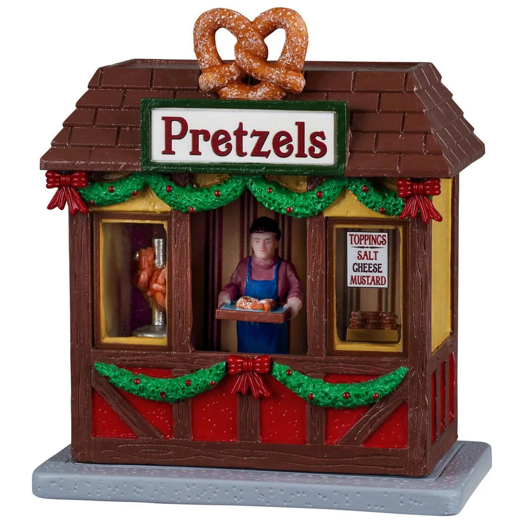 This image shows what this Lemax table accent looks like on a white background. It was designed to look like a wooden booth that sells pretzels. Under the roof and under the counter is decorated with garland and red bows. There's a large, fake pretzel on the roof. There's a guy behind the counter holding a tray of fresh pretzels. There's a sign in the window that reads "Toppings: Salt, Cheese, Mustard."