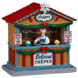 This image shows what this Lemax table accent looks like on a white background. It was designed to look like a wooden booth that sells crepes. The roof is red with a blue and yellow trim. There's also a white snowflake on the roof. Underneath the counter, the booth is decorated with green garland and red bows. There's a person behind the counter about to crack an egg. There are signs that read "Drinks: Coffee, Tea, Wine, Juice" and "Fillings: Chocolate, Caramel, Jam, Preserves, Fruit."