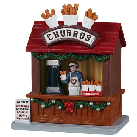 This image shows what this Lemax table accent looks like on a white background. It was designed to look like a wooden booth that sells churros. The roof is red and decorated with three large bouquets of fake churros. Underneath the counter, the booth is decorated with green garland, silver bells and red bows. There's a man behind the counter showing off a cup of freshly baked churros and wearing a white apron. There's a sign on the ground that reads "Menu: Cinnamon, Chocolate, Caramel, Eggnog, Gingerbread."