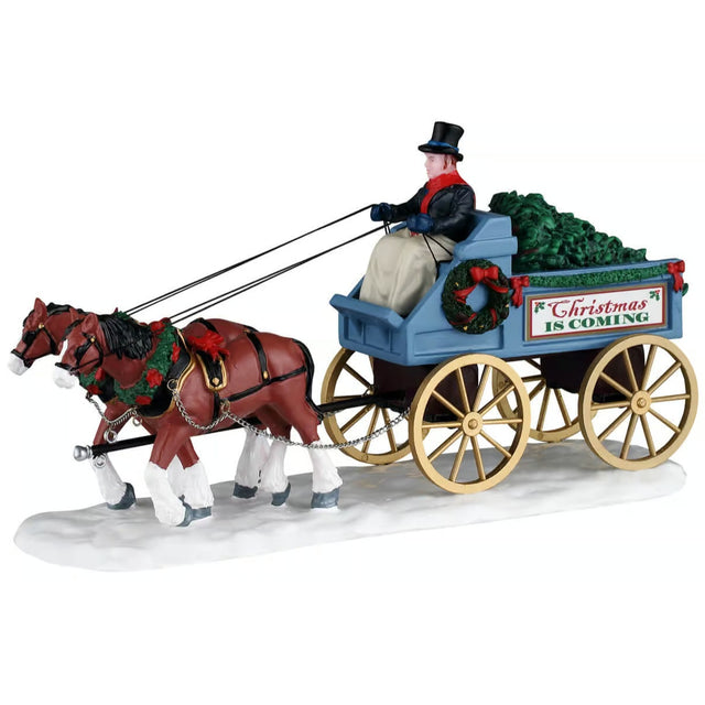 This image shows what this Lemax Caddington Village table accent looks like on a white background. It was designed to look like two horses pulling a large carriage through the snow. Inside the carriage, there are Christmas trees. There's a man steering the horses. He's wearing a black top hat, a black jacket and a tan colored blanket.