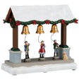 This image shows what this Lemax table accent looks like on a white background. It was designed to look like a wooden stall with three gold bells. The roof of the stall is covered in snow and garland decorated with red bows. There are three kids ringing the bells while a dog looks on.