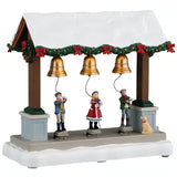 This image shows what this Lemax table accent looks like on a white background. It was designed to look like a wooden stall with three gold bells. The roof of the stall is covered in snow and garland decorated with red bows. There are three kids ringing the bells while a dog looks on.
