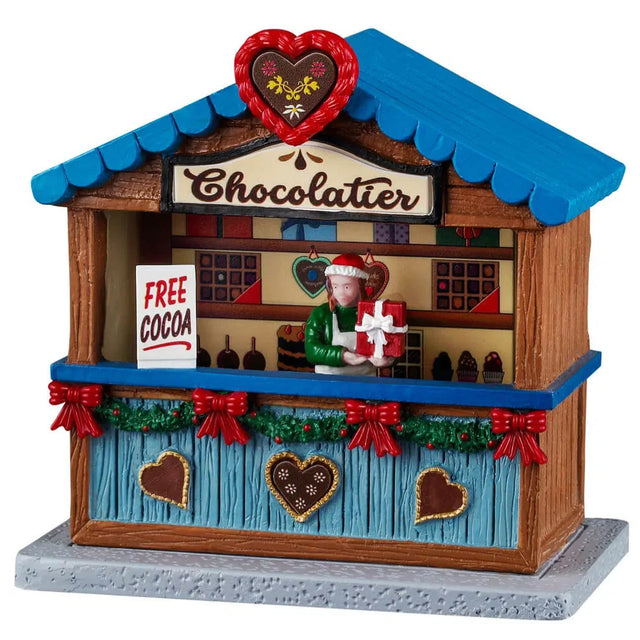 This image shows what this Lemax table accent looks like on a white background. It was designed to look like a wooden booth that sells chocolates. The roof and the counter are blue. Underneath the counter, the booth is decorated with green garland and red bows. The sign at the top of the booth reads "Chocolatier" and the sign on the counter reads "Free Cocoa." There's a woman behind the counter showing off a newly wrapped box of chocolates.