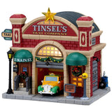 This image shows what this Lemax building looks like on a white background. It was designed to look like a red barn turned into a garage for taxis. There's a large, gold star on the roof. There are decorative garlands and wreaths. Inside the garage, there's a yellow and green colored taxi.