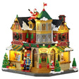 This image shows what this Lemax building looks like on a white background. It was designed to look like a very large toy shop. There's a large fake Santa sitting on the roof, waving. The roof and sidewalk are covered in snow. There are Christmas decorations all over, as well as some nutcrackers standing guard by the front door.