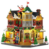 This image shows what this Lemax building looks like on a white background. It was designed to look like a very large toy shop. There's a large fake Santa sitting on the roof, waving. The roof and sidewalk are covered in snow. There are Christmas decorations all over, as well as some nutcrackers standing guard by the front door.