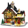 This image shows what this Lemax Caddington Village lighted building looks like on a white background. It was designed to look like a house turned into an art studio. The roof and the sidewalk has snow on it. Inside the window, you can see a woman painting. There's a decorated tree, a bouquet of flowers, an easel with a painting on it and a snowman on the sidewalk.
