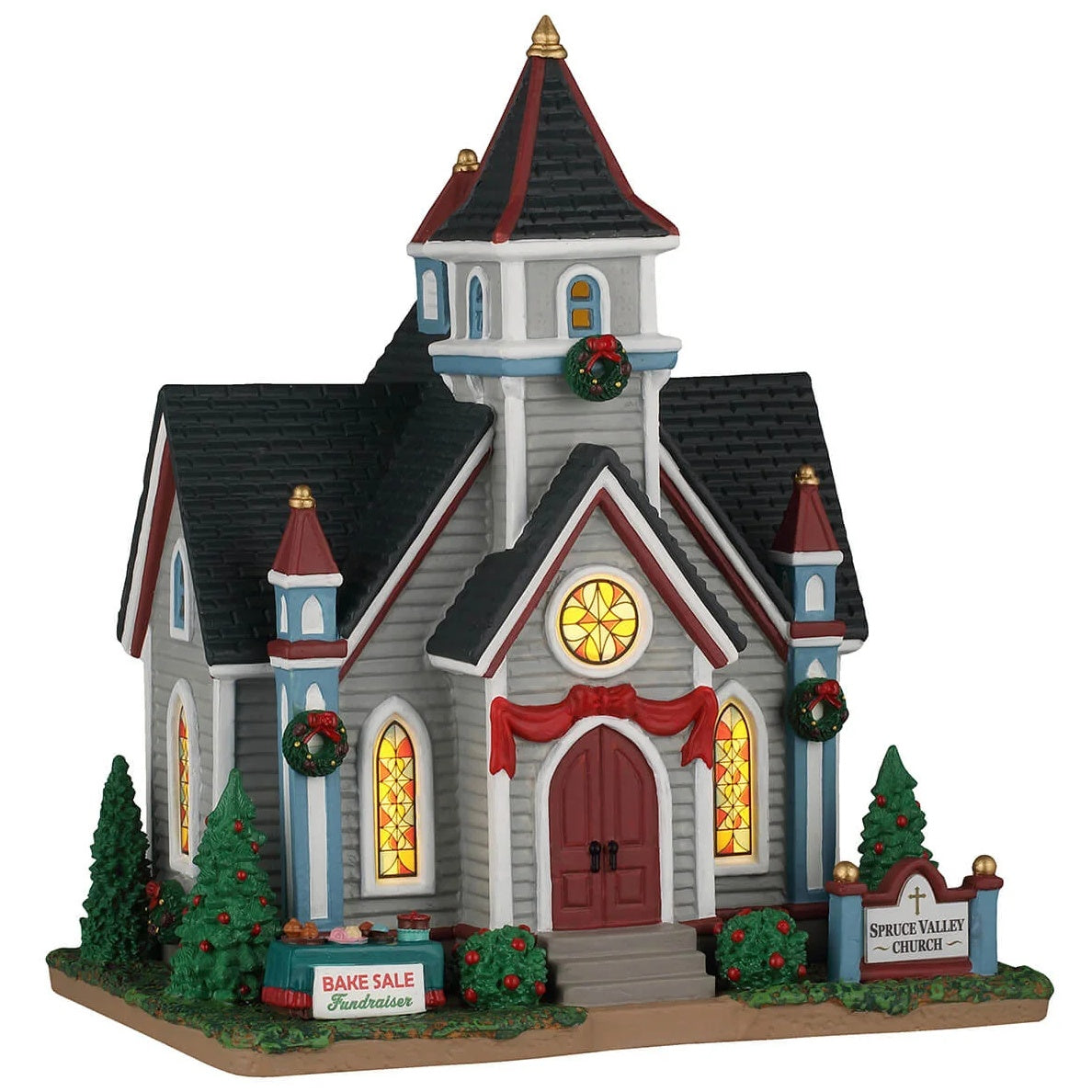 This image shows what this Lemax building looks like on a white background. It was designed to look like a large chapel. The chapel is decorated with red bows and wreaths. There are three stained glass windows. There's a sign on a table that reads "Bake Sale Fundraiser."