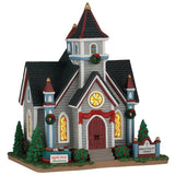 This image shows what this Lemax building looks like on a white background. It was designed to look like a large chapel. The chapel is decorated with red bows and wreaths. There are three stained glass windows. There's a sign on a table that reads "Bake Sale Fundraiser."