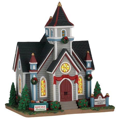 This image shows what this Lemax building looks like on a white background. It was designed to look like a large chapel. The chapel is decorated with red bows and wreaths. There are three stained glass windows. There's a sign on a table that reads "Bake Sale Fundraiser."