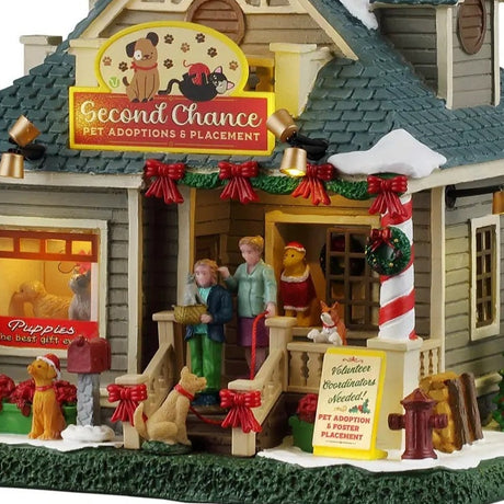 This is an up close view of the lighted building. You can see there's a dog wearing a Santa hat near the mailbox and there's a dog on a red leash standing on the front stairs. The sign near the fire hydrant reads "Volunteer Coordinators Needed! Pet Adoption & Foster Placement."