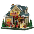 This image shows what this Lemax Caddington Village lighted building looks like on a white background. It was designed to look like a house turned into a pet adoption agency. The roof is covered in snow. There are people and dogs standing on the front porch. Inside the window, you can see puppies playing. Around the house, there are decorated trees, presents, chopped wood and a red fire hydrant. The stairs and the roof are decorated with red bows and garlands.