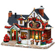 This image shows what this Lemax building looks like on a white background. It was designed to look like a bed and breakfast house. The roofline is covered in a poinsettia garland and lights. There are tables outside and people are decorating them with poinsettias.