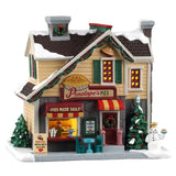 This image shows what this Lemax building looks like on a white background. It was designed to look like a house turned into a pie shop. The snow and the sidewalk are covered in snow. There's a walk-up counter with a person behind the counter serving pies. There's a table near the front door, along with a snowman holding a pie.