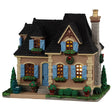 This image shows what this Lemax building looks like on a white background. It was designed to look like a cheery cottage with lots of plants around the cottage. The window shutters and the doors are a light blue color. There are potted plants, bushes and trees. Outside the windows, there are planters of flowers.