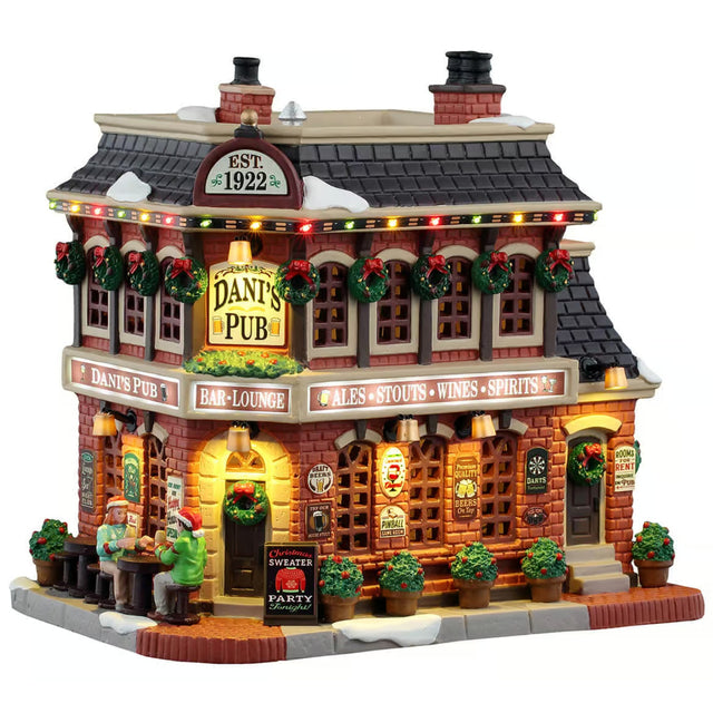 This image shows what this Lemax Caddington Village lighted building looks like on a white background. It was designed to look like a large, old-timey cottage turned into a pub. The windows are decorated with wreaths and the sidewalk is decorated with potted plants. There are two people eating at table on the sidewalk.