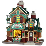 This image shows what this Lemax building looks like on a white background. It was designed to look like a tall cottage turned into a store. The roofs and the sidewalks are covered in snow. There are Christmas decorations on the building and near the front door.