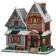 This image shows what this Lemax building looks like on a white background. It was designed to look like a snow-covered house. The left side of the house is blue and the right side of the house is red. There are red bows, garlands and wreath accents, as well as a bunch of wrapped presents on the front sidewalk.