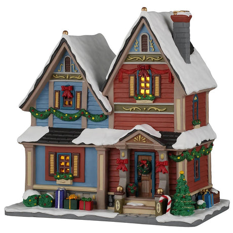 This image shows what this Lemax building looks like on a white background. It was designed to look like a snow-covered house. The left side of the house is blue and the right side of the house is red. There are red bows, garlands and wreath accents, as well as a bunch of wrapped presents on the front sidewalk.