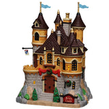 This image shows what this Lemax building looks like on a white background. It was designed to look like a large castle. There are blue flags on the steeples and decorated trees and wreaths. There's a sign near the front gate that reads "Arie's Castle."