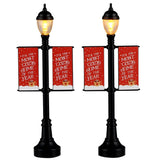 This image shows what these two Lemax Village accessories look like on a white background. They were designed to look like lamp posts decorated with banners for winter. The lamp posts are black. The banners re red and read "It's the Most Cozy Time of the Year."