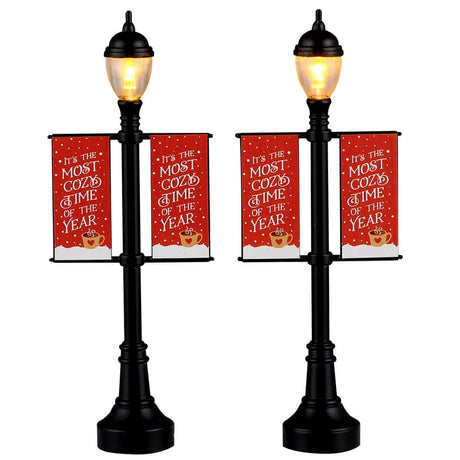 This image shows what these two Lemax Village accessories look like on a white background. They were designed to look like lamp posts decorated with banners for winter. The lamp posts are black. The banners re red and read "It's the Most Cozy Time of the Year."