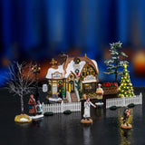 This is a lifestyle image showing this Lemax house set on a fake background. It was designed to look like an old-timey cottage turned into a bakery. There are three trees, one of them is lighted. There's a mom and a young daughter out for a walk in their winter's best. There's also a baker chasing down a young boy who stole some bread.