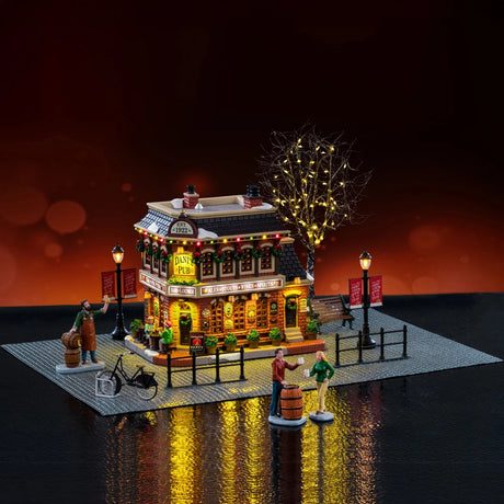This image shows what this Lemax house set looks like with a fake background. The set is designed to look like a tall pub on a stone landscape. There are fences and lampposts and people standing around wooden kegs. There's a tree with lights on it behind the house.