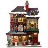 This image shows what this Lemax building looks like on a white background. It was designed to look like a tall shopping center made up of different shops. There's snow on the roof and on the sidewalk. There are nutcrackers, decorated wreaths and garlands and wrapped presents all over. Inside the window, you can see a bunch of different children's toys on display, like a rocking horse and a giant teddy bear dressed like Santa.