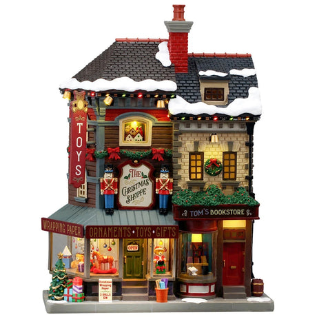 This image shows what this Lemax building looks like on a white background. It was designed to look like a tall shopping center made up of different shops. There's snow on the roof and on the sidewalk. There are nutcrackers, decorated wreaths and garlands and wrapped presents all over. Inside the window, you can see a bunch of different children's toys on display, like a rocking horse and a giant teddy bear dressed like Santa.