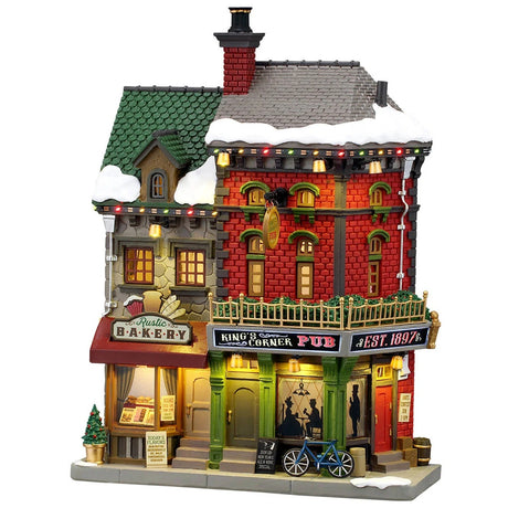 This image shows what this Lemax building looks like on a white background. It was designed to look like a tall building consisting of two business. The left is called the Rustic Bakery and inside the windows, you can see the different menus. The right is called King's Corner Pub. There's snow on the roof and on the sidewalk. 