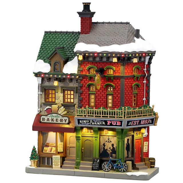 This image shows what this Lemax building looks like on a white background. It was designed to look like a tall building consisting of two business. The left is called the Rustic Bakery and inside the windows, you can see the different menus. The right is called King's Corner Pub. There's snow on the roof and on the sidewalk. 