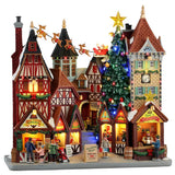 This image shows what this Lemax Caddington Village building facade looks like on a white background. It was designed to look like a strip mall made up of old-timey, Victorian-era-type buildings. They're all decorated for Christmas with garlands. There's a giant decorated Christmas tree with Santa and his reindeer flying high above the building.