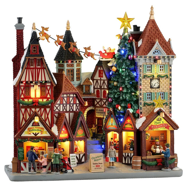 This image shows what this Lemax Caddington Village building facade looks like on a white background. It was designed to look like a strip mall made up of old-timey, Victorian-era-type buildings. They're all decorated for Christmas with garlands. There's a giant decorated Christmas tree with Santa and his reindeer flying high above the building.